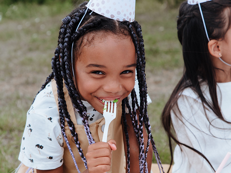 A girl wearing a party hat eating cake and grinning