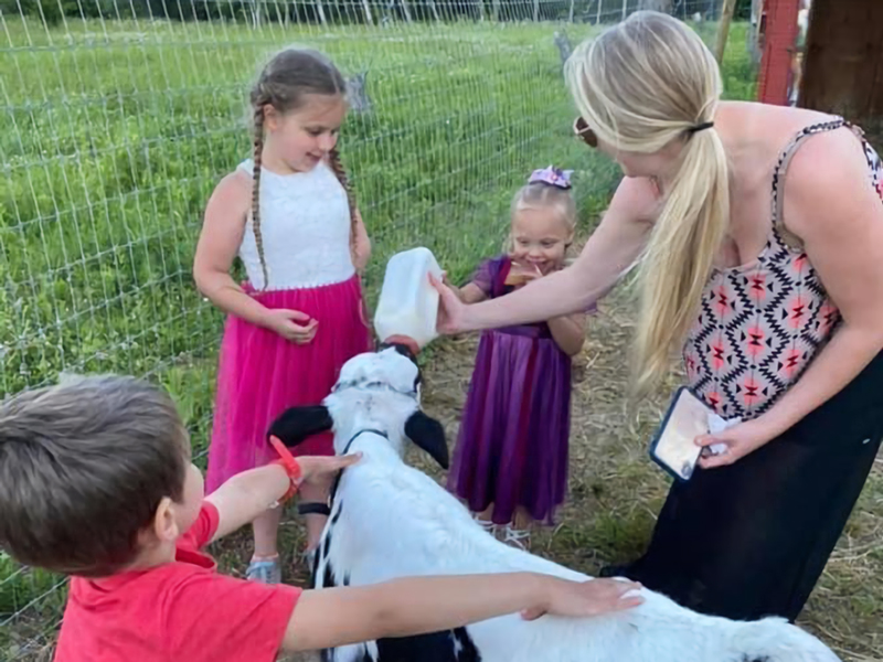 Three smiling kids, two girls in dresses and a boy in a red shirt, gathered around a woman bottle-feeding a calf. The boy is holding the calf.
