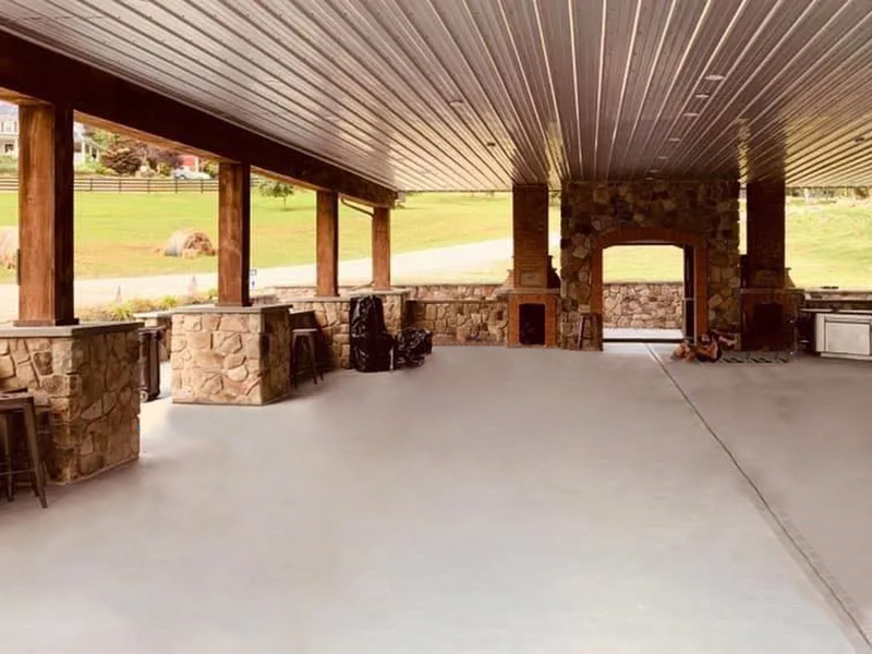 The outdoor pavilion, a sunny space with a cement floor and metal ceiling, supported by stone and wood columns, surrounded by grass